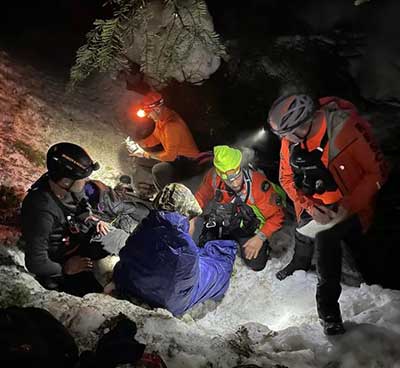 Rescuers in helmets gather around a search subject at night with headlamps lit and snow on the ground