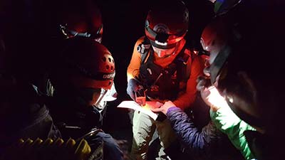 Several search and rescue people huddle in the dark with helmets and headlamps with a GPS unit looking at a map