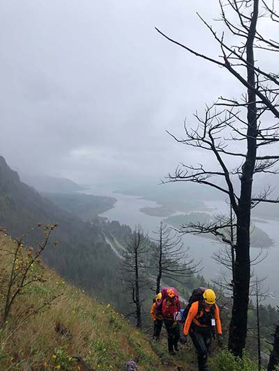 Three people wearing helmets and backpacking gear walk a trail along a hill with mist and a river in the background