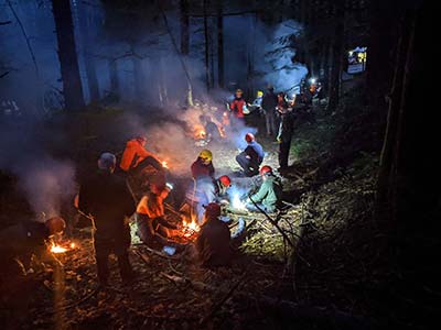 Multiple search and rescue people with gear sit among campfires in the dark with smoke rising around them