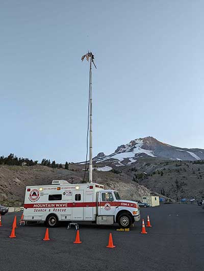 A large truck with Mountain Wave Search and Rescue on the side with a red stripe, a mountain in the distance, and a large antenna extending from the truck