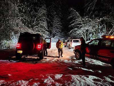 Multiple search and rescue vehicles in the snow with snow covered trees and a person walking in the middle of the snowy road