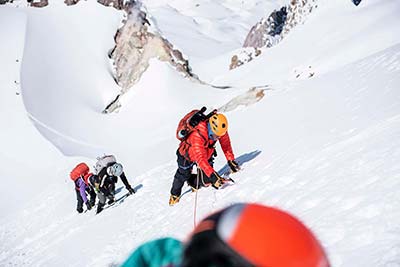 Three people in winter gear are roped and ascending a snow covered mountain slope with a helmet of another person in the foreground