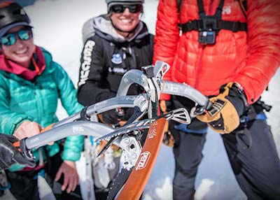 Three people in mountain climbing gear hold their ice axes together in the middle of the photo