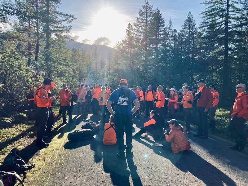 Several search and rescue volunteers, mostly wearing orange and blue, gathered together on a campground road in the forest